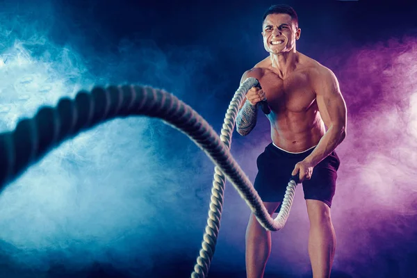 El bodybulder atlético que mira trabaja hacia fuera con la cuerda de batalla en fondo oscuro del estudio con el humo. Fuerza y motivación — Foto de Stock