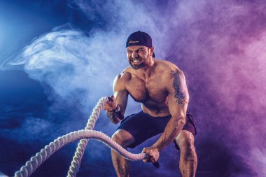 Sakallı, atletik görünümlü vücut bulucu. Karanlık stüdyo arka planında savaş halatıyla dumanla çalışıyor. Kuvvet ve motivasyon