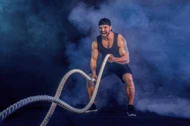 Sakallı, atletik görünümlü vücut bulucu. Karanlık stüdyo arka planında savaş halatıyla dumanla çalışıyor. Kuvvet ve motivasyon