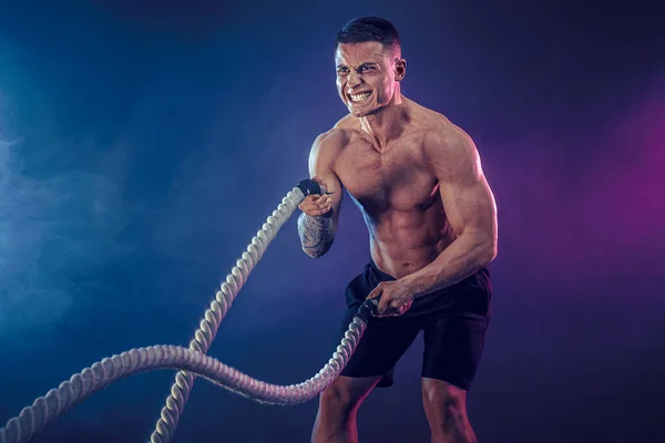 Athletic ser bodybulder träna med strids rep på mörk studio bakgrund med rök. Styrka och motivation — Stockfoto