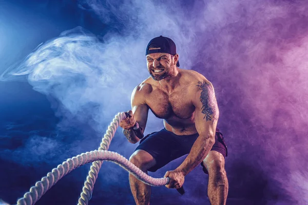 Sakallı, atletik görünümlü vücut bulucu. Karanlık stüdyo arka planında savaş halatıyla dumanla çalışıyor. Kuvvet ve motivasyon