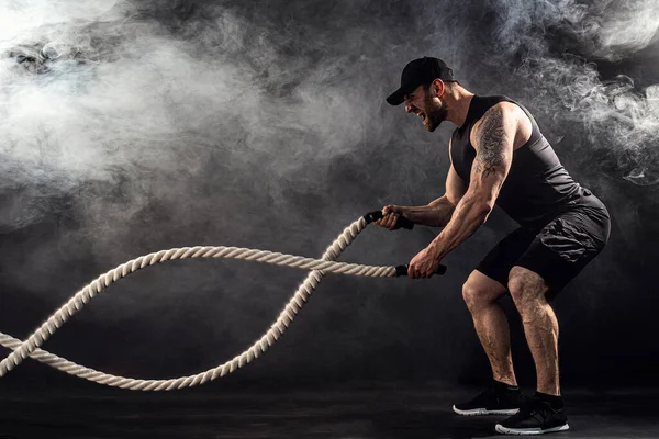Bearded athletic looking bodybulder work out with battle rope on dark studio background with smoke. Strength and motivation — Stock Photo, Image