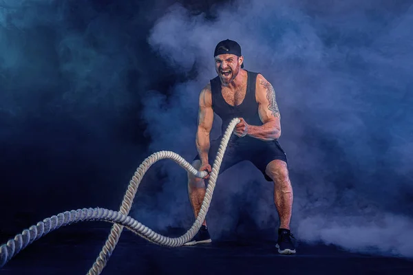 Atlético barbudo olhar bodybulder trabalhar com corda de batalha no fundo do estúdio escuro com fumaça. Força e motivação — Fotografia de Stock