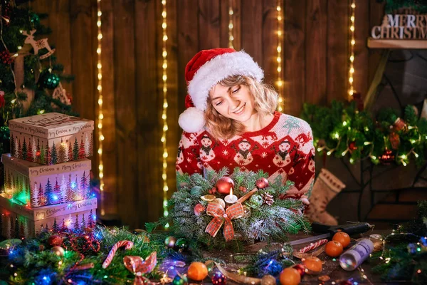 Жінка робить різдвяний вінок ручної роботи на столі. Різдвяні прикраси та композиції. Дівчина, одягнена в різдвяний дизайнерський светр, готує різдвяний вінок до свята — стокове фото