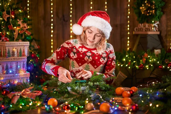 Mulher vestida com camisola de design de Natal fazendo grinalda de Natal artesanal na mesa para férias. Decoração e composição de Natal — Fotografia de Stock