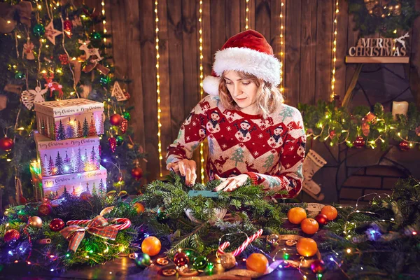 Жінка, одягнена в різдвяний дизайнерський светр, робить різдвяний вінок ручної роботи на столі для свята. Різдвяні прикраси та композиції — стокове фото