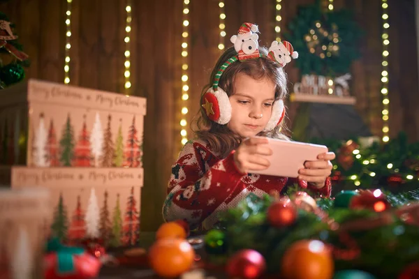 Flicka använder telefon nära julgran i den dekorativa interiören. Jul och nyår foto. — Stockfoto