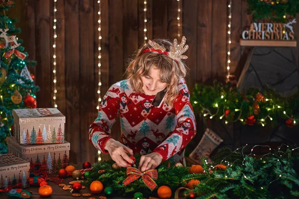 Жінка, одягнена в різдвяний дизайнерський светр, робить різдвяний вінок ручної роботи на столі для свята. Різдвяні прикраси та композиції — стокове фото