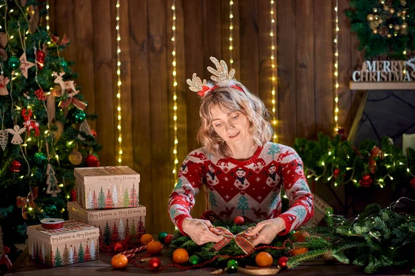 Donna vestita con maglione di design natalizio che fa ghirlanda di Natale fatta a mano sul tavolo per le vacanze. Decorazione e composizione natalizia — Foto Stock