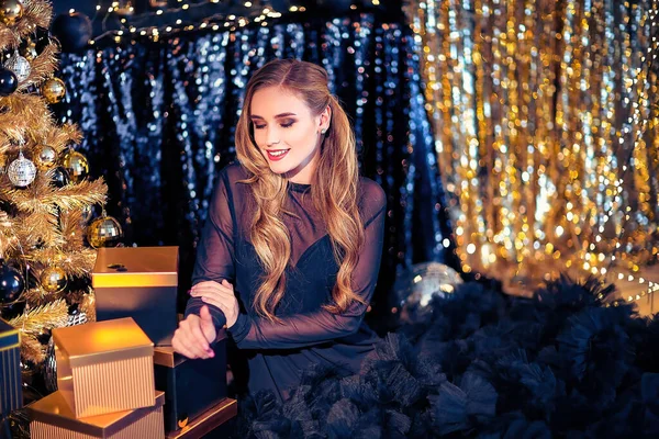 Jovem mulher bonita em elegante vestido de noite sentado no chão perto da árvore de Natal e apresenta em uma véspera de ano novo. Interior com decorações de Natal. DOF rasa e fundo bokeh — Fotografia de Stock