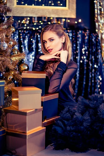 Νεαρή όμορφη γυναίκα με κομψό βραδινό φόρεμα κάθεται στο πάτωμα κοντά στο χριστουγεννιάτικο δέντρο και παρουσιάζει την παραμονή της Πρωτοχρονιάς. Εσωτερικό με χριστουγεννιάτικα στολίδια. Ρηχό DOF και bokeh φόντο — Φωτογραφία Αρχείου