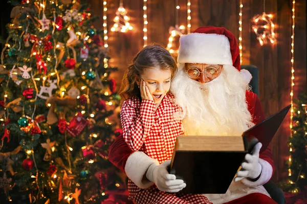 Weihnachtsmann öffnet und liest Zauberbuch mit kleinem niedlichen Mädchen im Pyjama — Stockfoto