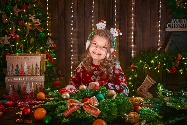 Mała dziewczynka dekorowanie wieniec iglasty w pobliżu choinki w dekoracyjnym wnętrzu. Boże Narodzenie i Nowy Rok zdjęcie — Zdjęcie stockowe
