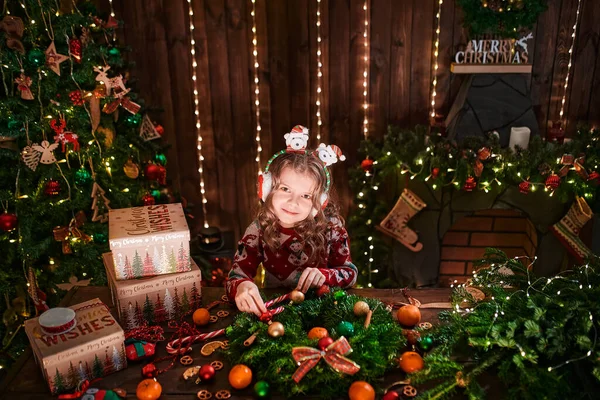 Маленька дівчинка прикрашає хвойний вінок біля ялинки в декоративному інтер'єрі. Різдво і Новий рік фото — стокове фото