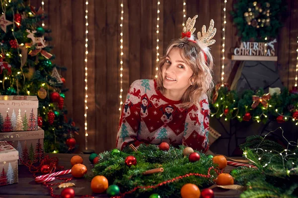 Mulher vestida com camisola de design de Natal, aro de chifre de veado feito à mão grinalda de Natal na mesa para férias, olhando para a câmera sorrindo. Decoração e composição de Natal — Fotografia de Stock