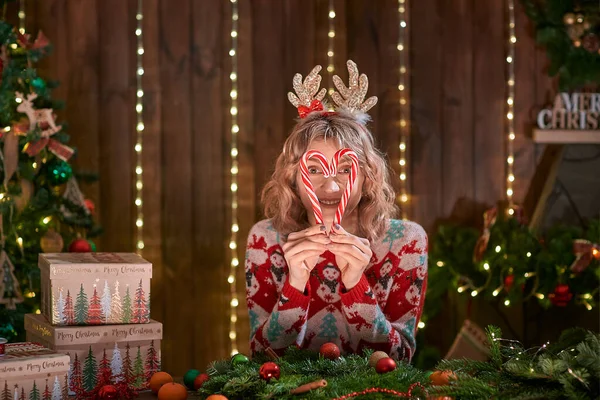 Жінка, одягнена в різдвяний светр, обруч з кільцем для оленів, робить різдвяний вінок ручної роботи на столі для відпочинку, робить обличчя, розважається, позує з різдвяними цукерками . — стокове фото