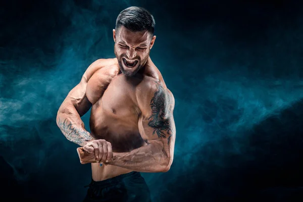 Hermoso poder atlético hombre culturista. Fitness cuerpo muscular sobre fondo de humo oscuro. . — Foto de Stock