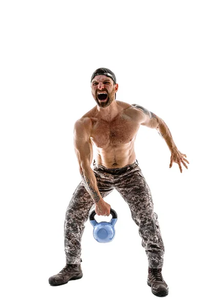 Atleta muscolare uomo bodybuilder in pantaloni mimetici con un allenamento nudo del busto con kettlebell su sfondo bianco. — Foto Stock