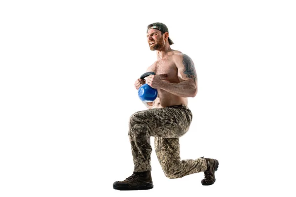 Gespierde atleet bodybuilder man in camouflage broek met een naakte romp workout met kettlebell op een witte achtergrond. — Stockfoto