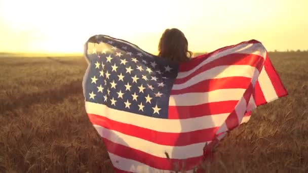 Menina de vestido branco vestindo uma bandeira americana enquanto corre em um belo campo de trigo — Vídeo de Stock