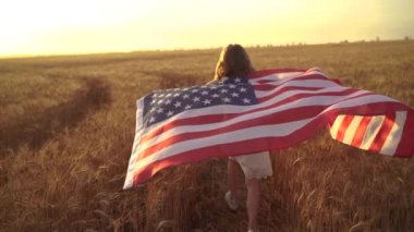 Beyaz elbiseli kız güzel bir buğday tarlasında koşarken Amerikan bayrağı takıyor.