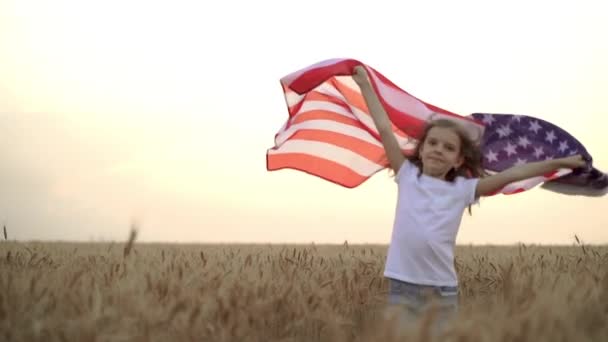 A kislány amerikai zászlót visel, miközben egy gyönyörű búzamezőn fut naplementekor. — Stock videók
