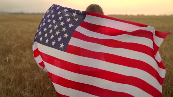A kislány amerikai zászlót visel, miközben egy gyönyörű búzamezőn fut naplementekor. — Stock videók