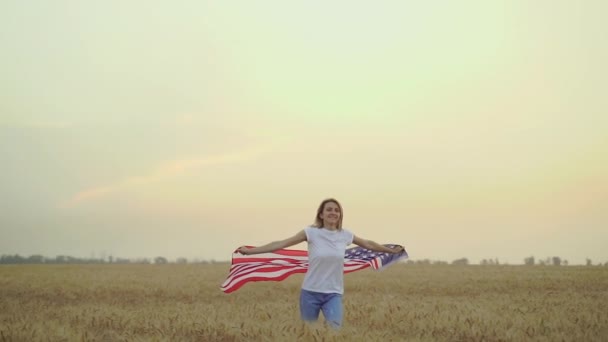 Женщина бежит и прыгает беззаботно с распростертыми объятиями над пшеничным полем, держа флаг США — стоковое видео
