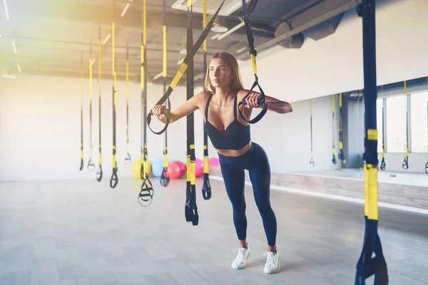 Beautiful young woman training with suspension trainer sling or suspension straps in gym. Upper body exercise concept on TRX