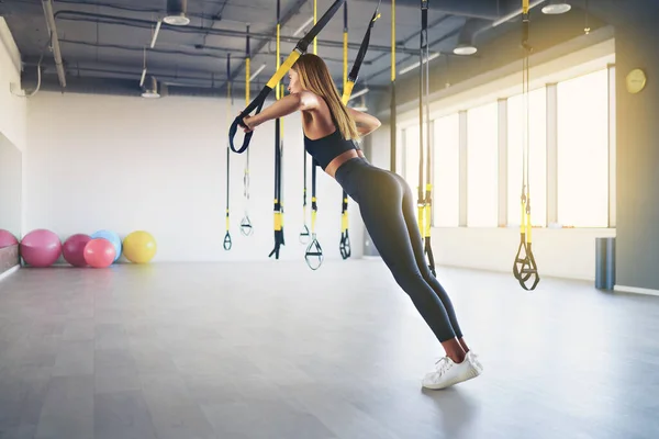 Beautiful young woman training with suspension trainer sling or suspension straps in gym. Upper body exercise concept on TRX