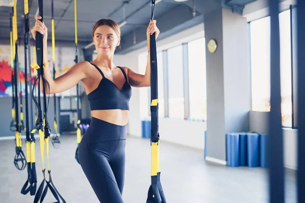 Beautiful young woman posing with suspension trainer sling or suspension straps in gym