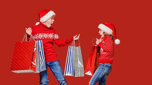Cute little girl in Santa hat hanging shopping bag with Christmas gifts of astonished boy during holiday celebration against red background — Stock Photo, Image