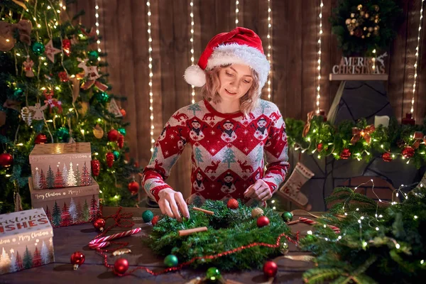 Mulher vestida com camisola de design de Natal fazendo grinalda artesanal na mesa para férias. Decoração e composição de Natal — Fotografia de Stock