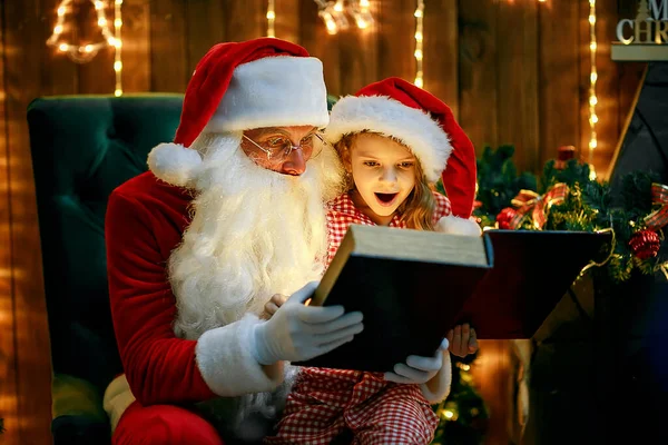 Weihnachtsmann öffnet und liest Zauberbuch mit kleinem niedlichen Mädchen im Pyjama — Stockfoto