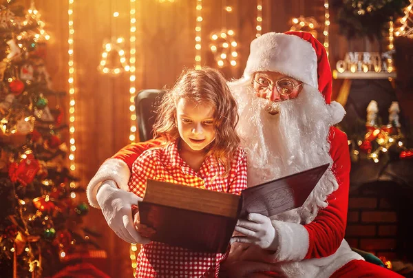 Jultomten öppna och läsa magiska bok med liten söt förvånad flicka i pyjamas — Stockfoto