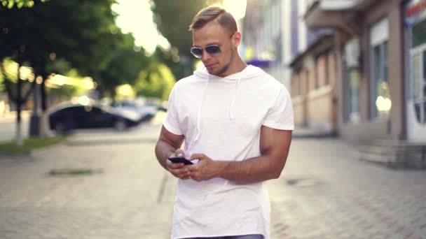 Jóképű, boldog férfi gépel a mobiltelefonján, miközben jó hangulatban áll a szabadban. Kaukázusi vidám fiatalember sms-ezés és koppintás okostelefon az utcán — Stock videók