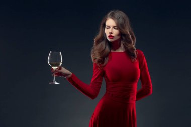 Parlak makyajlı, saç stili, kırmızı elbiseli, koyu arkaplanda bir cam ile poz veren bir kadın.