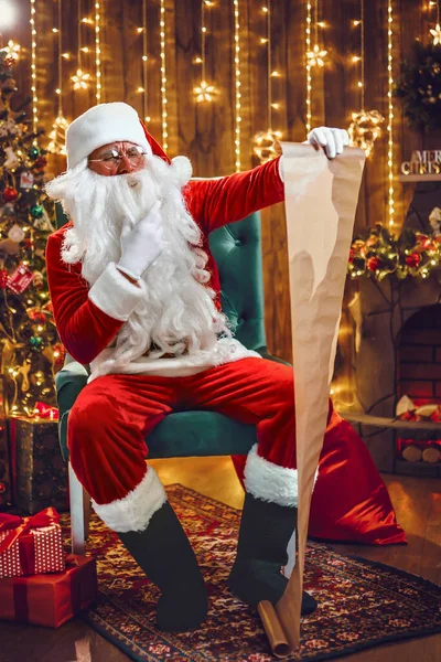 Porträt des Weihnachtsmannes, der zu Hause in der Nähe des Weihnachtsbaums in seinem Zimmer sitzt und Weihnachtsbrief oder Wunschzettel liest — Stockfoto