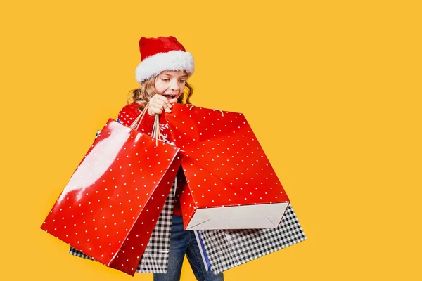 Adorable niño de Santa sombrero rojo y suéter con ciervos llevando coloridas bolsas de compras con regalos de Navidad sobre fondo amarillo — Foto de Stock