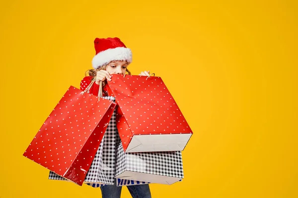 Adorable niño de Santa sombrero rojo y suéter con ciervos llevando coloridas bolsas de compras con regalos de Navidad sobre fondo amarillo — Foto de Stock