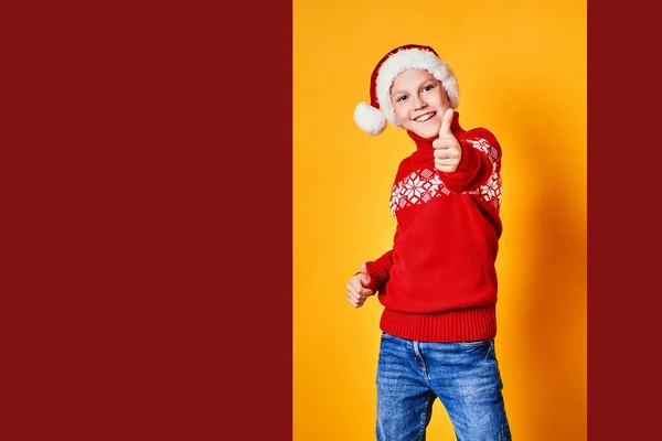 Niño en el sombrero de Santa y suéter de diseño de Navidad roja divertirse y mostrar el pulgar hacia arriba en el día de Navidad contra el fondo amarillo —  Fotos de Stock