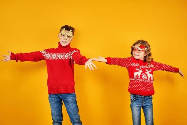 Дети в теплых красных рождественских свитерах и украшенных очках смотрят на камеру на желтом фоне — стоковое фото