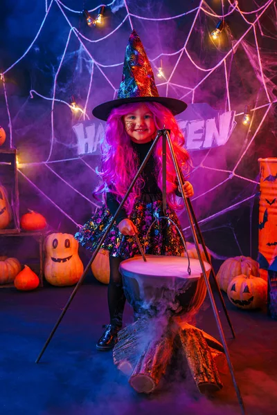 Malá krásná dívka v kostýmu čarodějnice slaví Happy Halloween party vařením lektvar v kouřící kádi, interiér s dýněmi — Stock fotografie