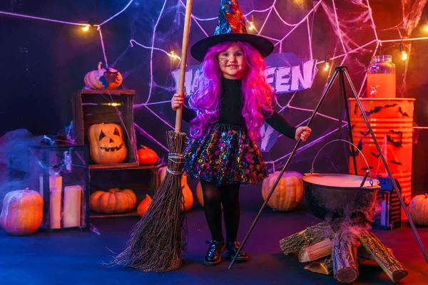 Kleine schöne Mädchen in einem Hexenkostüm feiert Happy Halloween Party, indem sie einen Trank in einem dampfenden Bottich brauen, Interieur mit Kürbissen — Stockfoto
