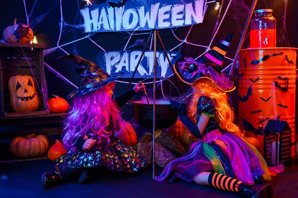 Dvě krásné holčičky v kostýmu čarodějnice slaví Happy Halloween party vařením lektvaru v kouřící nádrži, interiér s dýněmi — Stock fotografie