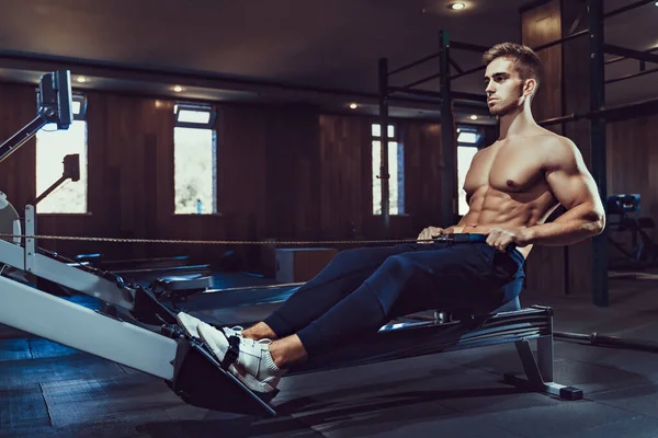 Muscular culturista en el entrenamiento de ropa deportiva de nuevo sentado en la máquina de ejercicio. Vista lateral del hombre con cuerpo de edificio del cuerpo perfecto en el gimnasio en la atmósfera oscura. Concepto de culturismo, fitness —  Fotos de Stock