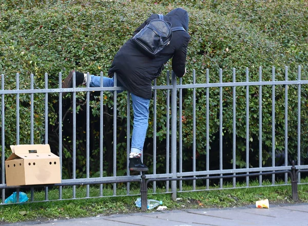 Bir erkekte bir ceket bir kukuleta ile düşük bir metal çitin üzerinden tırmanıyor