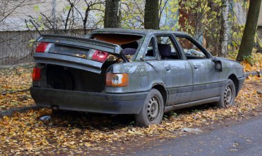 Bir apartmanın bahçesindeki sarı yaprakların üzerinde terk edilmiş eski, koyu gri bir araba.