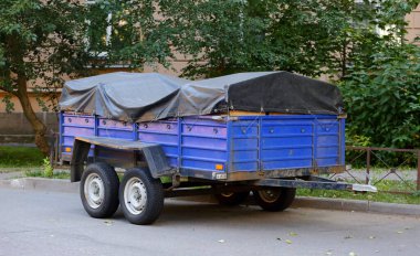 Koyu tenteli dört tekerlekli ağır bir kargo karavanı yeşil ağaçların yanına park edilmiş.