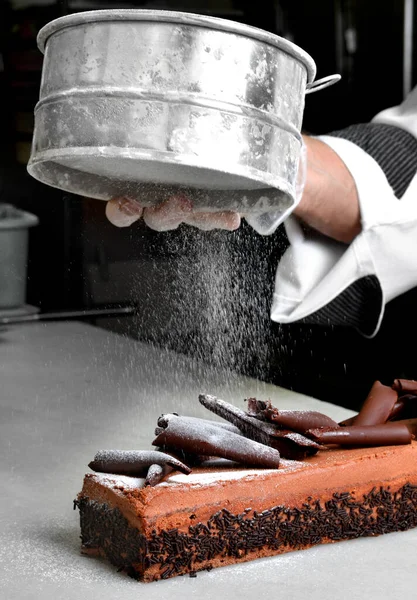 The pastry chef\'s hand with a strainer distributes icing sugar on the chocolate cake